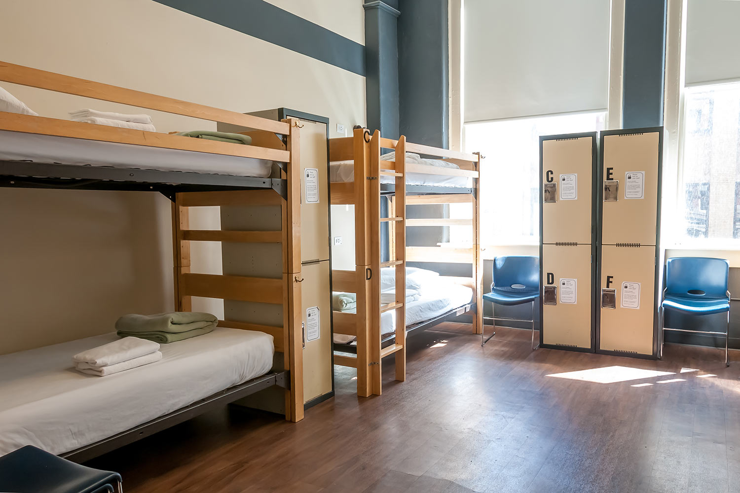 a small dorm room at HI Chicago hostel with two sets of bunk beds and four secure lockers for guests' belongings. There are two large windows with blinds that can be adjusted to let in light.
