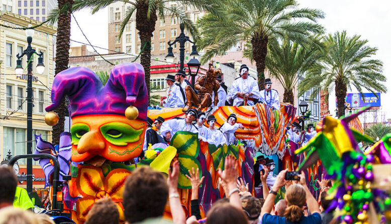 a colorful float goes down the street in New Orleans during Mardi Gras, surrounded by palm trees and an excited crowd