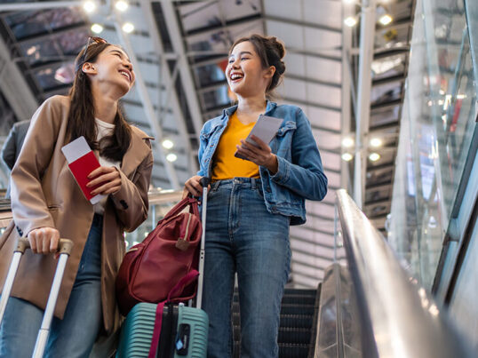 Two young women on an escalator at the airport with suitcases and passports