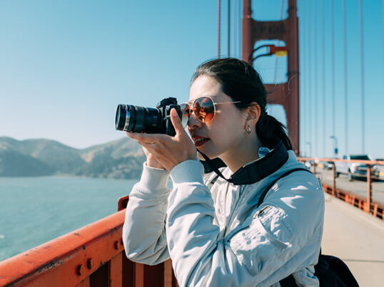 a young woman in sunglasses stands on the golden gate bridge in san francisco taking a photo with a camera with an extended lens