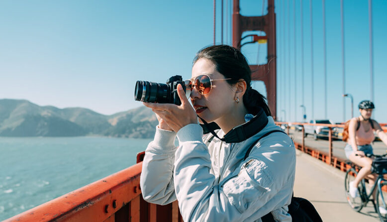 a young woman in sunglasses stands on the golden gate bridge in san francisco taking a photo with a camera with an extended lens