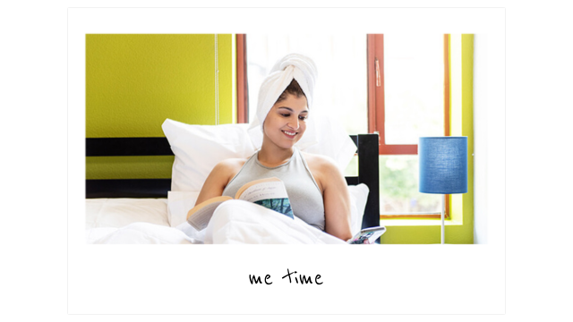 a young traveler lays in a full-sized bed with clean white linens reading and looking at her phone. She has a towel wrapped around her wet hair.