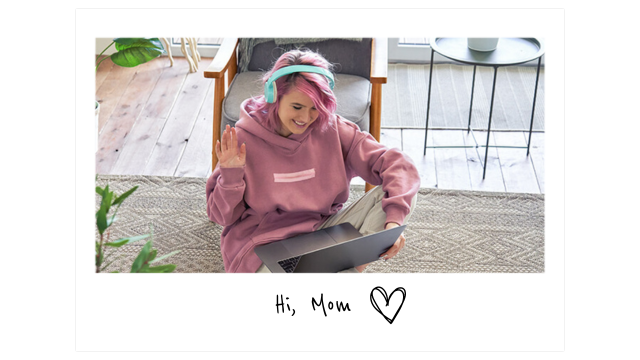 a young woman with pink hair and a pink sweatshirt and blue headphones sits on the floor with an open laptop waving at the screen