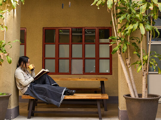 a young woman relaxes at a picnic table outside while reading a book and sipping from a yellow mug.