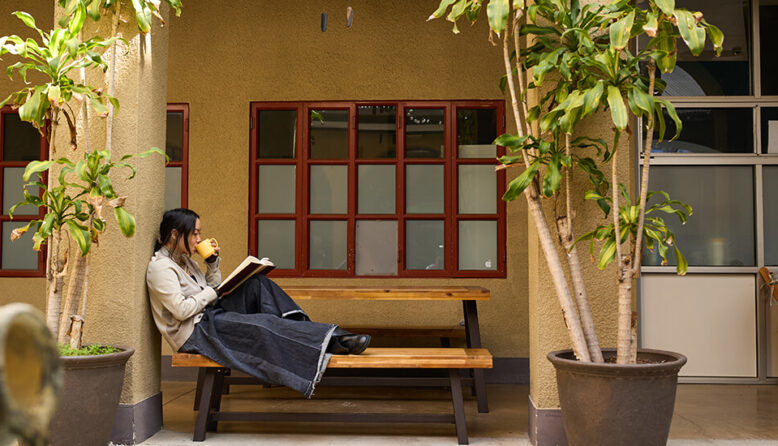 a young woman relaxes at a picnic table outside while reading a book and sipping from a yellow mug.