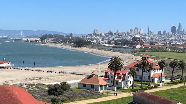 An overhead view of Crissy Field in San Francisco shows a stretch of sandy beach along the bay, with the San Francisco city skyline in the background.