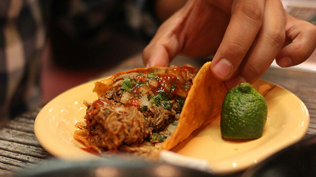 a hand picking up a birria taco, which sits on a small yellow plate next to a wedge of lime.