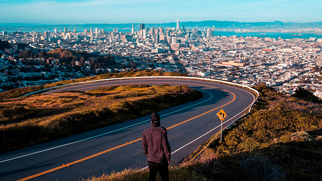 a man with his back to the camera looks out over San Francisco from Twin Peaks with a winding road in front of him