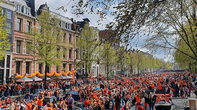 A crowd people dressed in orange fill up a street alongside beautiful houses in Amsterdam on Kings Day