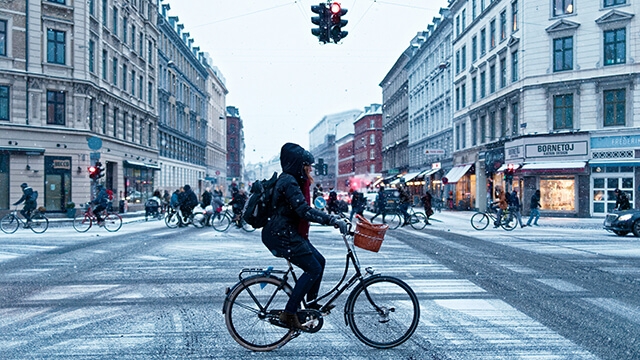 a person in a winter hat and coat rides a bicycle down a street in copenhagen dusted with snow