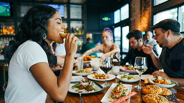 A group of young friends sit at a long table covered in plates of food enjoying a meal together